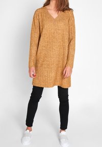 Senapsgul ribbad stickad tunika klänning med V-ringning, långa ärmar, avslappnad passform, kombinerad med svarta leggings och vita slip-on skor.