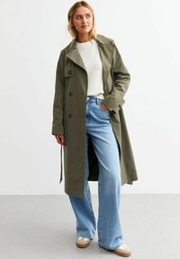 Olivgrön trenchcoat med dubbelknäppning, knappdetaljer och bälte, bärs över en ljus tröja och vida blå jeans.
