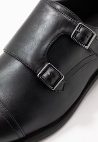 Chaussure en cuir noir avec une texture lisse, dotée de deux boucles métalliques ajustables et de détails cousus sur la partie supérieure.