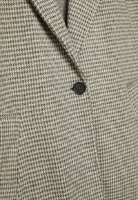Blejzer s houndstooth vzorom v sivi in bež barvi. Narejen iz teksturirane tkanine, z črnim gumbom in izrezom na ovratniku.