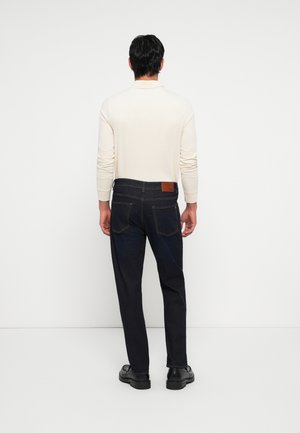 Mann trägt cremefarbenes Langarmshirt, dunkelblaue Jeans mit sichtbaren Gesäßtaschen und schwarze Schuhe, steht vor einer weißen Wand.