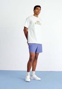 Pilkas trumpomis rankovėmis marškinėliai su spalvinga „New Balance“ grafika, derinami su šviesiai purpurinėmis šortais ir baltais sportbačiais ant mėlynos grindų.