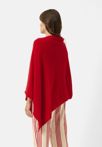 Maglione poncho rosso realizzato in tessuto morbido. Presenta un design drappeggiato con un collo alto e un orlo triangolare. Indossato sopra pantaloni a righe beige e rossi.