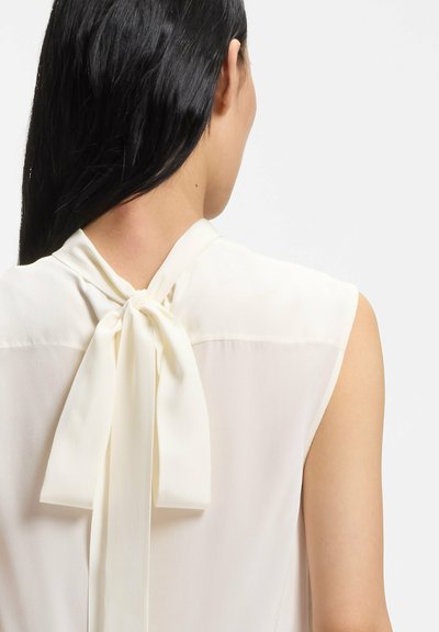 Blusa blanca sin mangas con cierre de lazo en la parte trasera. Tejido liso, ligero y de corte relajado. Diseño simple y elegante.