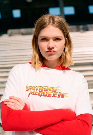 Young woman with blonde hair, arms crossed, wearing a white "Lightning McQueen" T-shirt over a red long-sleeve shirt, standing in front of stairs.
