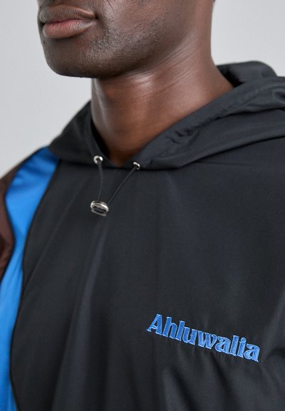 Svart huva jacka med blå detaljer, dragkedja och broderad "Ahluwalia" logotyp. Materialet har en jämn, glansig textur.