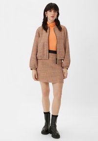 Karierter Bomberjacke mit elastischen Bündchen, orangefarbener Rollkragenpullover und einem passenden Minirock. Schwarze Knöchelstiefel setzen einen Kontrast zum Outfit.