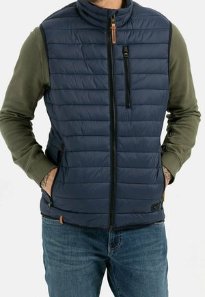 Mann trägt eine marineblaue gesteppte Weste mit Front- und Brusttaschen mit Reißverschluss über einem olivfarbenen Langarmshirt und blauen Jeans, die Hände in den Taschen der Weste.