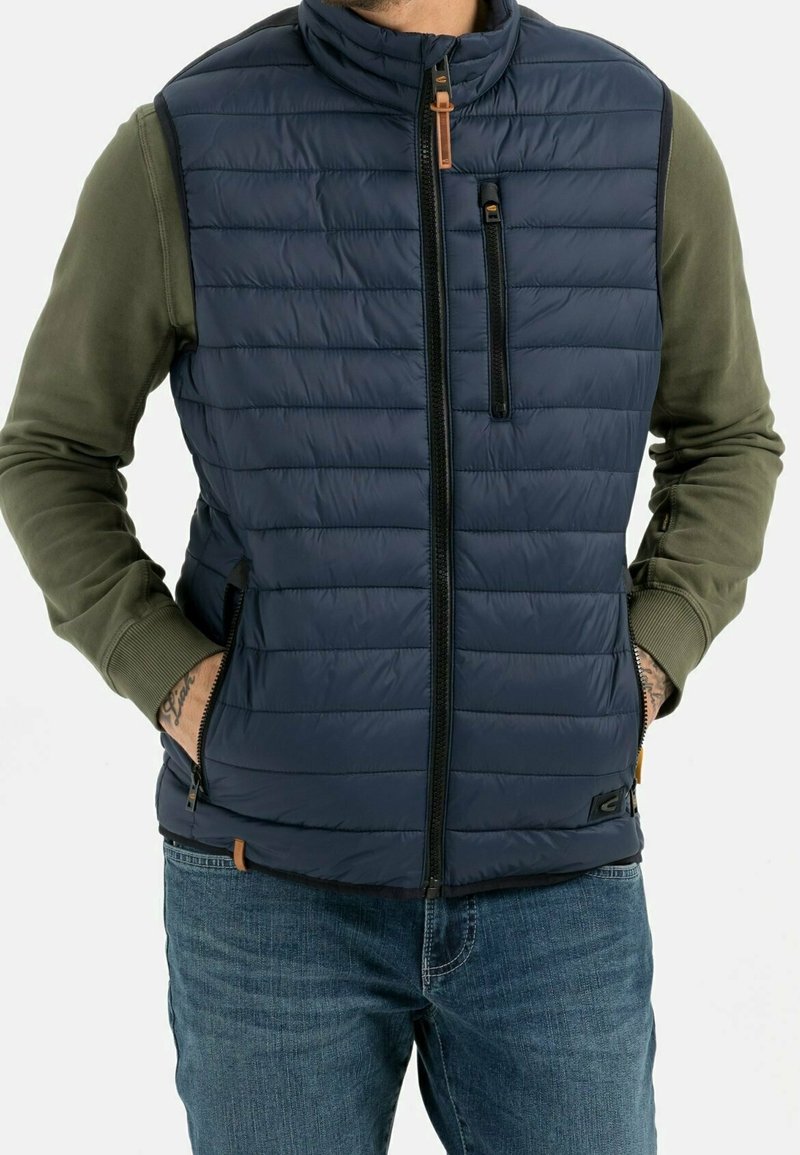 Homme portant une veste matelassée bleu marine avec poches zippées à l'avant et sur la poitrine, par-dessus une chemise olive à manches longues et un jean bleu, les mains dans les poches de la veste.
