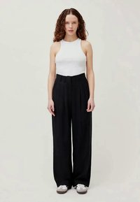 Mujer con cabello rizado, vistiendo una blusa blanca sin mangas, pantalones de pierna ancha negros de alta cintura y zapatillas blancas, de pie contra un fondo liso.