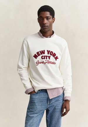 Jonge man draagt een witte trui met rode ""New York City Gant Athletic"" tekst, daaronder een gestreept hemd en een blauwe spijkerbroek, staand en ontspannen.