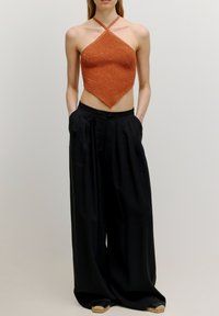 Oranges Halftertop mit einer strukturierten, glitzernden Oberfläche und einer diamantförmigen Form. Kombiniert mit lockeren schwarzen Hosen, die Falten und Taschen besitzen.