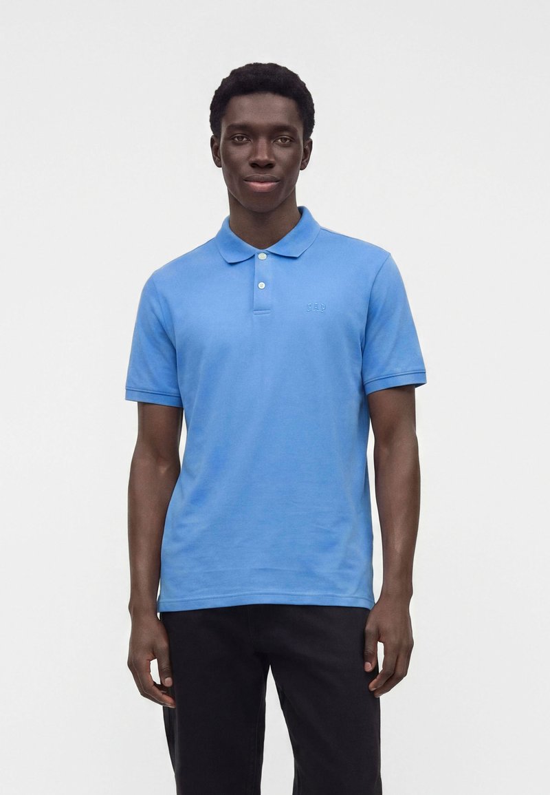 Homme debout devant un fond uni, portant un polo bleu clair à manches courtes et un pantalon noir, face à l’avant, les mains détendues.