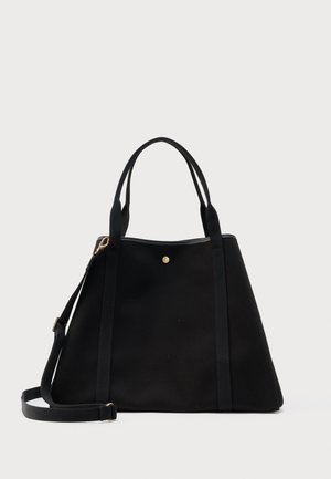 Sac fourre-tout trapézoïdal noir avec deux bretelles d'épaule, une sangle bandoulière amovible, et un petit détail bouton doré sur un fond uni.