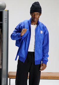 Zila bombjera jaka ar teksturētu rakstu, rāvējslēdzēja aizdari un logotipa detaļu. Kombinēta ar melnām biksēm un zilu mugursomu. Uz galvas adīta cepure.