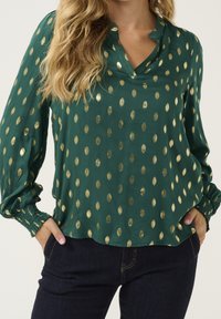 Blusa verde con pois a forma di foglia dorata, vestibilità ampia, scollo a V, maniche lunghe con polsini elastici, texture liscia. Abbinata a jeans scuri.