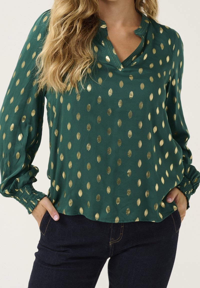 Blusa verde con pois a forma di foglia dorata, vestibilità ampia, scollo a V, maniche lunghe con polsini elastici, texture liscia. Abbinata a jeans scuri.