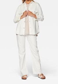 Biała koszula z kordurowej tkaniny z dwoma kieszeniami na piersi, noszona na jasnobeżowej bluzce, w zestawie z białymi spodniami kordurowymi i beżowymi sandałami na klamrę.