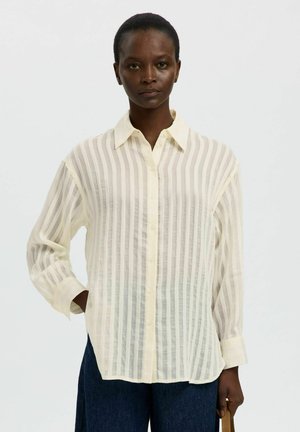 Femme à la peau foncée portant une chemise blanche transparente rayée à manches longues et ample, boutonnière, et un pantalon foncé, tenant une sangle marron dans une main.