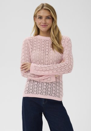 Vrouw met lang blond haar, die een roze gebreide trui met kantpatroon en donkerblauwe spijkerbroek draagt, met gekruiste armen en een lichte glimlach.