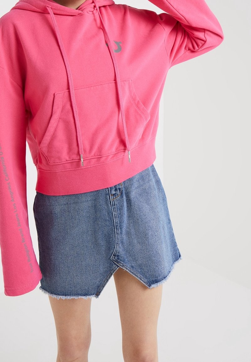 Sudadera corta rosa con bolsillo frontal y detalle de cordón, combinada con una falda de mezclilla azul con dobladillo deshilachado y corte en ángulo.