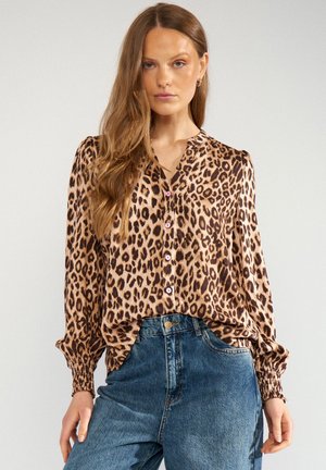 Frau mit langen Haaren, die eine Leopardenmuster-Bluse und blaue hoch taillierte Jeans trägt, steht vor einem schlichten, hellen Hintergrund.