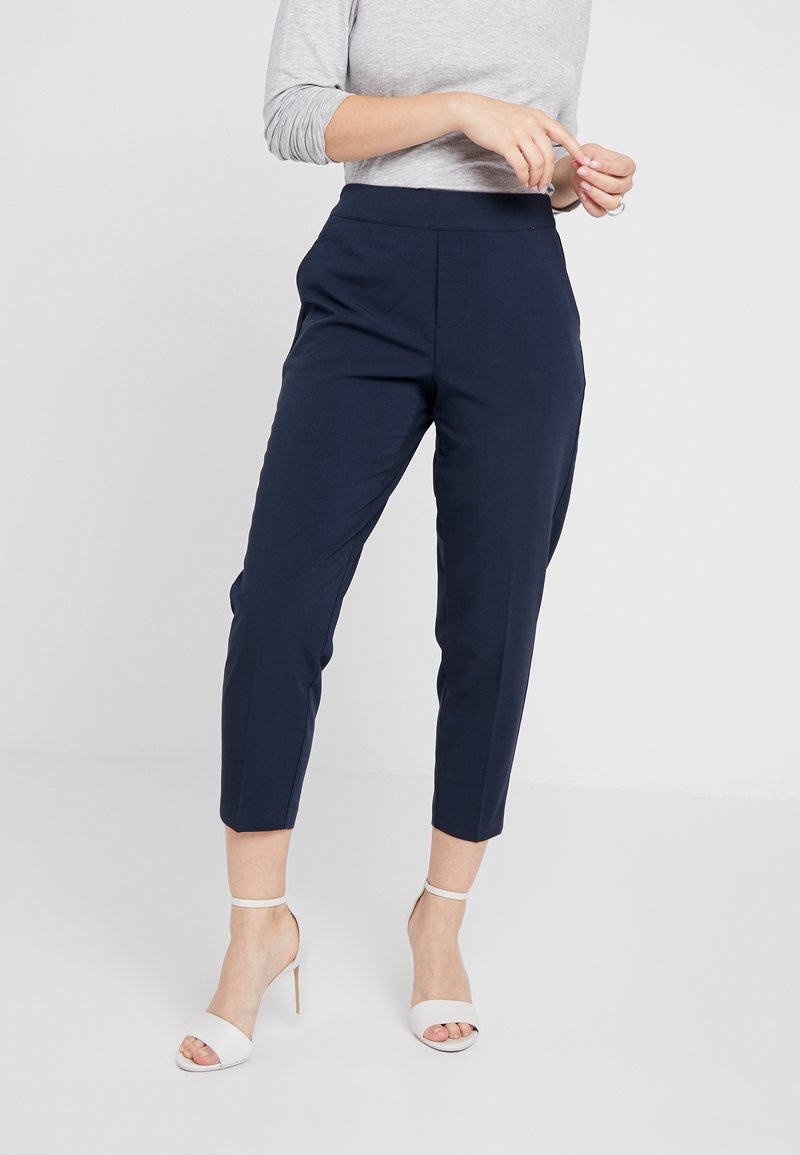 Pantalon court bleu marine en tissu lisse, avec une jambe fuselée et des plis sur le devant. Porté avec des sandales blanches à talons et un haut gris clair.
