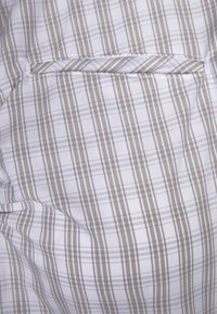 Vaaleanvärinen kangas, jossa valkoinen tausta ja ohuet ruskeat ja harmaat ruudulliset raidat. Pieni, vaakasuora taskudetalji mukana.