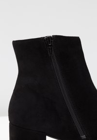 Stivale alla caviglia in suede nero con cerniera laterale e tacco a blocco su uno sfondo chiaro uniforme.