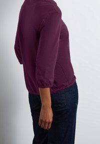 Person wearing a purple long-sleeve shirt and dark blue jeans, hand relaxed by their side, standing against a plain light background.