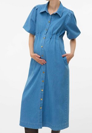 Femme enceinte portant une robe midi en jean bleu boutonnée, une main sur le ventre, l'autre main dans la poche, debout sur un fond blanc.