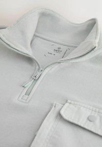 Lysegrå hættetrøje med lynlås og høj krave, har en frontlomme, ribbede manchetter og en lynlås med matchende trækkesnor. Størrelse M mærkat synlig.