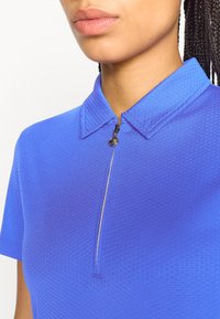 Zila polo krekls ar teksturētu audumu, īsām piedurknēm un ar rāvējslēdzēju priekšpusē. Funkcijas - apkakles dekoltē ar smalku ģeometrisku rakstu.