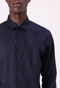 Camicia blu navy con bottoni, realizzata in cotone. Presenta un colletto a punta, una patta frontale con sei bottoni e una texture liscia.