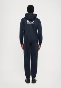 Survêtement navy comprenant une veste à capuche avec un logo EA7 blanc dans le dos et un pantalon ajusté assorti avec des poignets élastiques.