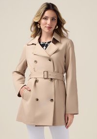 Beige dubbelzijdige trenchcoat met een riem in de taille, zijzakken en grote knopen, gedragen over een geblokte top. Soepele textuur en slim fit.