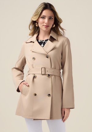 Beige doppelreihiger Trenchcoat mit tailliertem Gürtel, seitlichen Taschen und großen Knöpfen, getragen über einem gemusterten Oberteil. Glatte Textur und taillierte Passform.