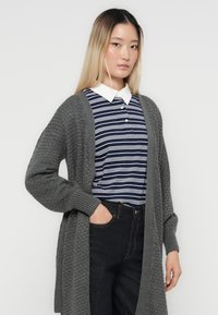 Grauer gestrickter Cardigan mit einem strukturierten Muster, getragen über einem navy-blauen und weißen gestreiften Poloshirt, kombiniert mit dunklen Jeans.