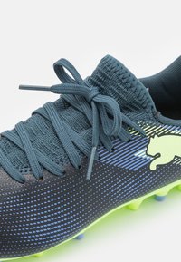 Scarpa da calcio con tomaia di un blu scuro e nero, superficie testurizzata, suola verde brillante e lacci blu tonali. Presenta un dettaglio del logo in evidenza.