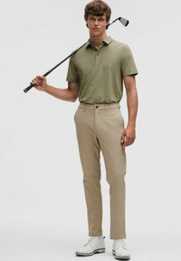 Męska oliwkowa koszulka polo z krótkimi rękawami, w zestawie z beżowymi spodniami i białymi butami golfowymi, trzymająca kij golfowy na neutralnym tle.