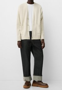 Krämsk knittröja med ribbade manschetter, V-hals och framlommor, lager på en vit t-shirt, kombinerad med mörka jeans och bruna skor.