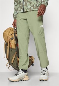 Pantaloni externe de un verde deschis, cu picioare înguste, având un logo pe piciorul inferior. Asortați cu un rucsac bej și pantofi albi.