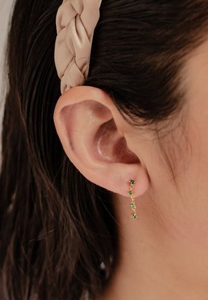 Goldene Ohrringe mit einem kaskadierenden Design und grünen Edelsteinen. Glänzende Oberfläche, zarte Kettenglieder und ein subtiler Glanz auf der Haut.