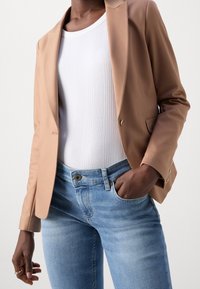 Blazer marron coupé sur mesure avec une finition lisse, porté sur un haut blanc côtelé et un jean ajusté bleu clair, avec des poches et un détail bouton.