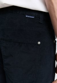 Granatowe sztruksowe spodnie z pionowym prążkowaniem, z tylnią kieszenią i widoczną niebieską metką marki. Z elementami ozdobnymi w kolorze srebrnym.