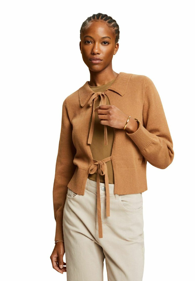 Cardigan en tricot marron avec un col, fermeture par lien et texture côtelée, porté sur un top vert olive, associé à un pantalon beige clair.