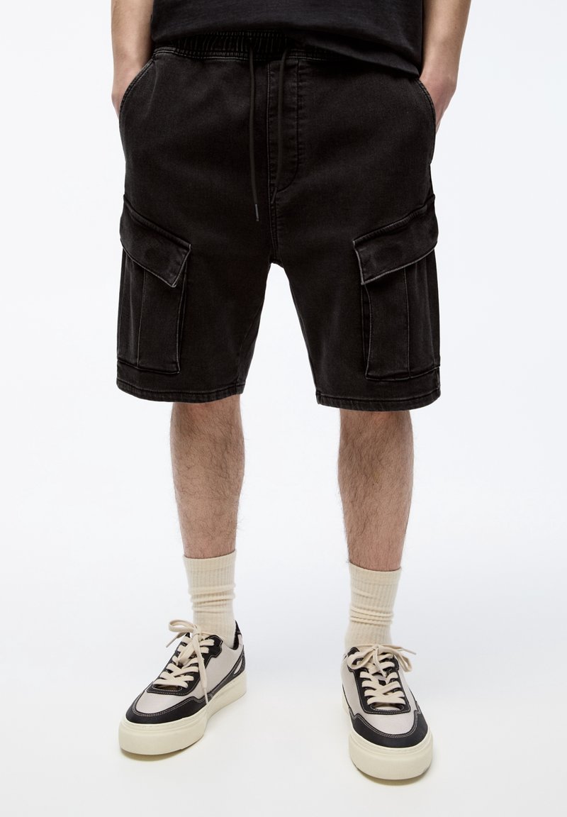 Shorts cargo en denim noir avec une ceinture élastique, deux poches latérales et deux poches cargo à l'avant. Portés avec des chaussettes courtes crème et des baskets.