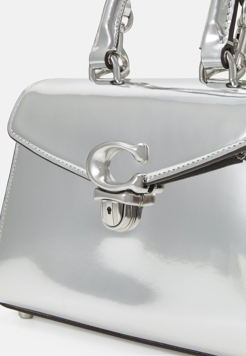 Sac à main en métal argenté avec un design structuré, doté d'un fermoir proéminent et de poignées élégantes. Texture lisse avec des détails de couture.
