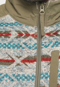 Veste en polaire avec un corps en tricot gris à motifs comportant des designs bleus, rouges et bruns ; accents unis vert olive ; fermeture éclair complète.