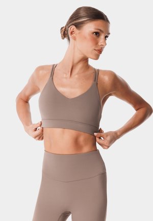 Wysportowana kobieta poprawia taupe sportowy biustonosz, nosi dopasowane legginsy z wysokim stanem, włosy związane w kok, na jasnym, jednolitym tle.